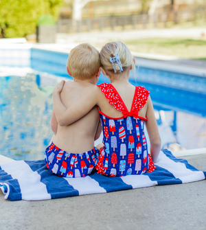 Boys and girl sitting poolside on a stripe towel wearing red, white and blue swimsuits, back