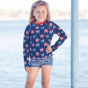 A young girl wearing a long sleeve rashguard in  Red,Wht,Blue Stars & navy Stripe swim shorts standing outside, lifestyle, front - Millieloveslily.com