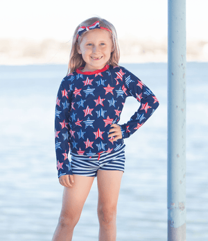 A young girl wearing a long sleeve rashguard in  Red,Wht,Blue Stars & navy Stripe swim shorts standing outside, lifestyle, front - Millieloveslily.com