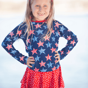 young girl wearing a  long sleeve  rashguard  in Red,Wht,Blue Stars & Skirty Bottom with red polka dots standing outside, lifestyle,front