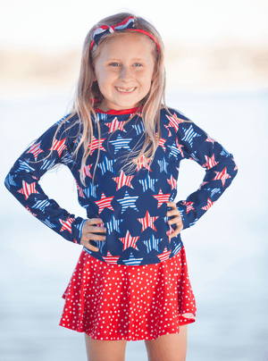 young girl wearing a  long sleeve  rashguard  in Red,Wht,Blue Stars & Skirty Bottom with red polka dots standing outside, lifestyle,front